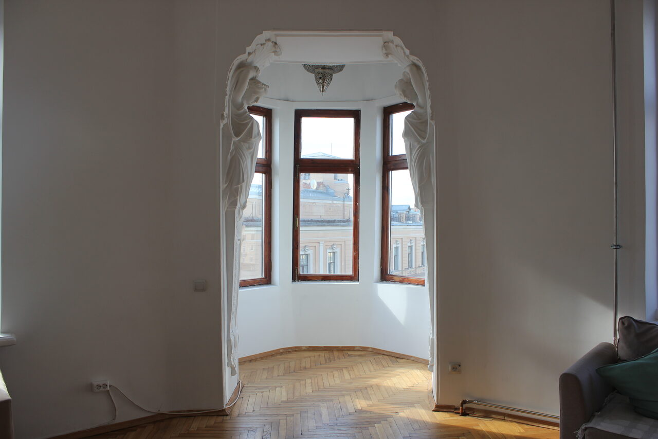 Арендовать Cassiopea (Кассиопея). Зона в светлой комнате с паркетом, декором в виде скульптур на арке; эркер с тремя окнами, вид на городские здания; справа часть дивана.