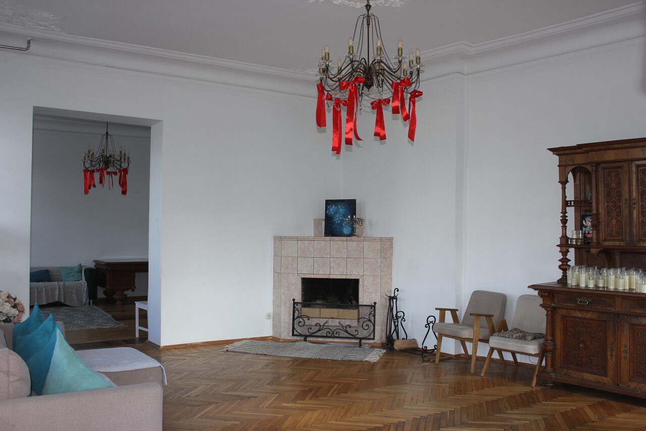 Арендовать Cassiopea (Кассиопея). Гостиная с паркетным полом и белыми стенами. В центре камин с декоративной решёткой, над ним картина. Справа деревянный буфет. Диван с цветными подушками, два кресла и столик. Две люстры с красными лентами. Освещение естественное.