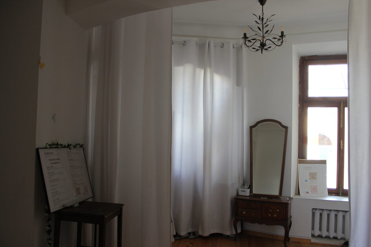 Арендовать Cassiopea (Кассиопея). Комната с деревянным полом, белыми занавесками, потолочным светильником, старинным зеркалом на комоде. На стене окно с деревянной рамой и радиатором. Справа от зеркала настольный мольберт с картиной. Слева столик с меню.