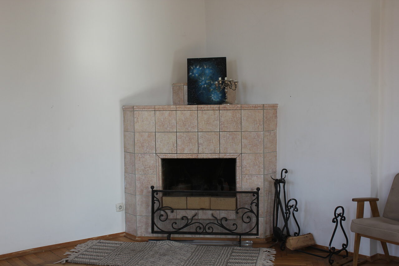 Арендовать Cassiopea (Кассиопея). Комната с камином, облицованным светлой плиткой. Вокруг него декоративная кованая решетка, рядом инструменты для камина. На камине картина и подсвечник. На полу светлый ковер, рядом кресло. Стены белые.