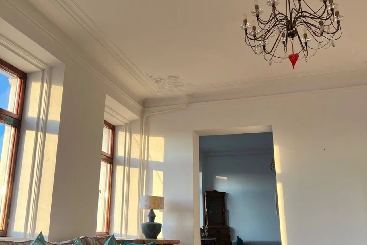 Арендовать Cassiopea (Кассиопея). Просторная комната с высокими потолками и белыми стенами. На потолке висит большая люстра. В комнате несколько окон с деревянными рамами, один торшер. На заднем плане виден другой отсек. Солнечный свет падает на стены.