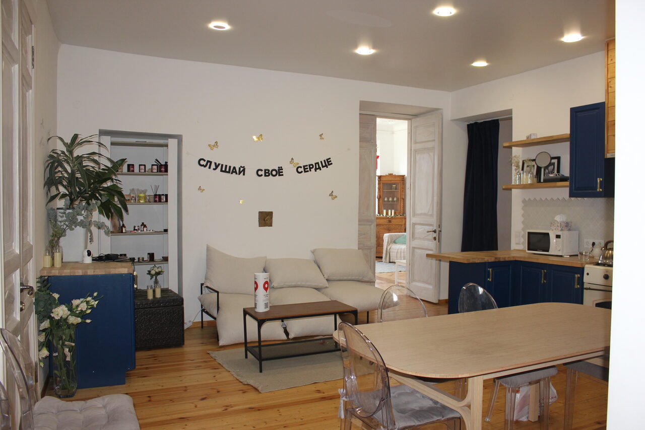 Арендовать Cassiopea (Кассиопея). Гостиная с кухонной зоной: деревянный пол, белые стены, потолочные светильники. Мягкий диван, кофейный столик, обеденный стол со стульями. Слева стеллаж с растениями. Кухня оснащена микроволновой печью и плитой. На стене надпись «Слушай своё сердце».