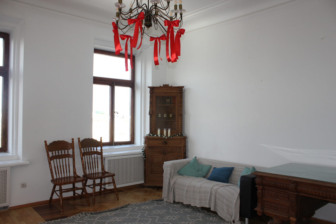 Арендовать Cassiopea (Кассиопея). Гостиная с деревянным паркетом, белыми стенами и большим окном. Мебель: два деревянных стула, диван с пледом и подушками, деревянный шкаф и письменный стол. На потолке люстра с красными лентами.