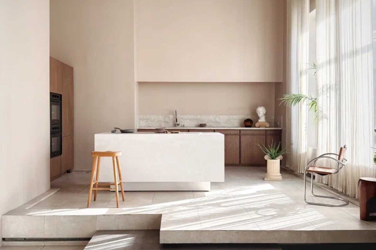 Арендовать Киото. Светлый зал с кухней. Современная кухня с открытой планировкой, белый остров с деревянным стулом, встроенный двойной шкаф-духовка, деревянные шкафы, мраморная столешница, светло-бежевые стены, большое окно с тюлевыми шторами, металлический и пластиковый стул, растения в горшках.
