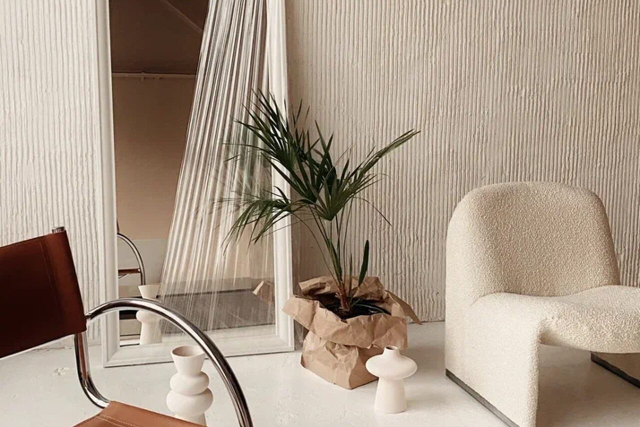 Арендовать Киото. Светлый зал с кухней. Минималистичное пространство с креслом белого цвета, округлой формы, растением в бумажной упаковке на полу, большим зеркалом у стены, кремовой настенной текстурой и элементами мебели из коричневой кожи и металла.