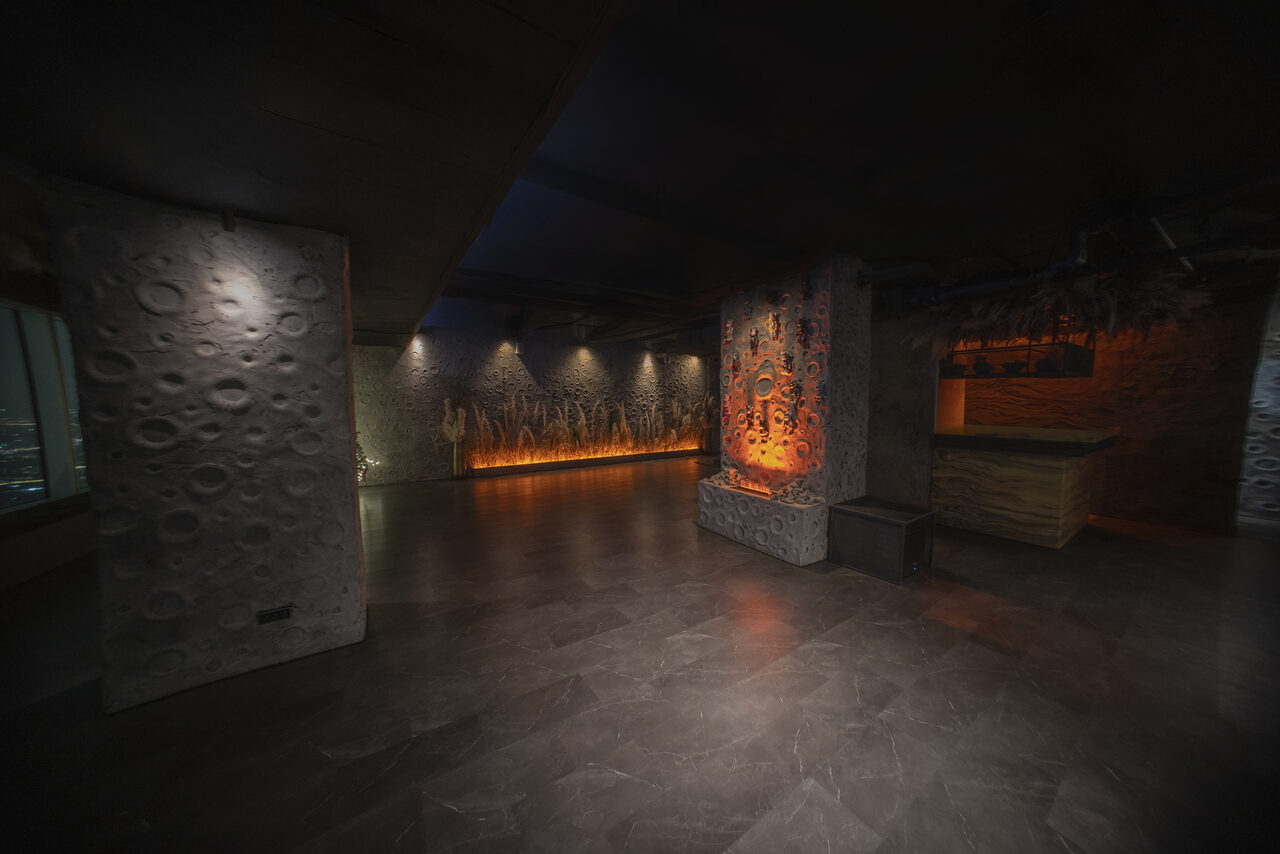 Арендовать ATMOSFERA 53(Атмосфера 53). Просторное помещение с тёмным полом и потолком, текстурированные стены с декоративной подсветкой. Стена с узорами и оранжевым освещением. Барная стойка справа. Мягкое рассеянное освещение.