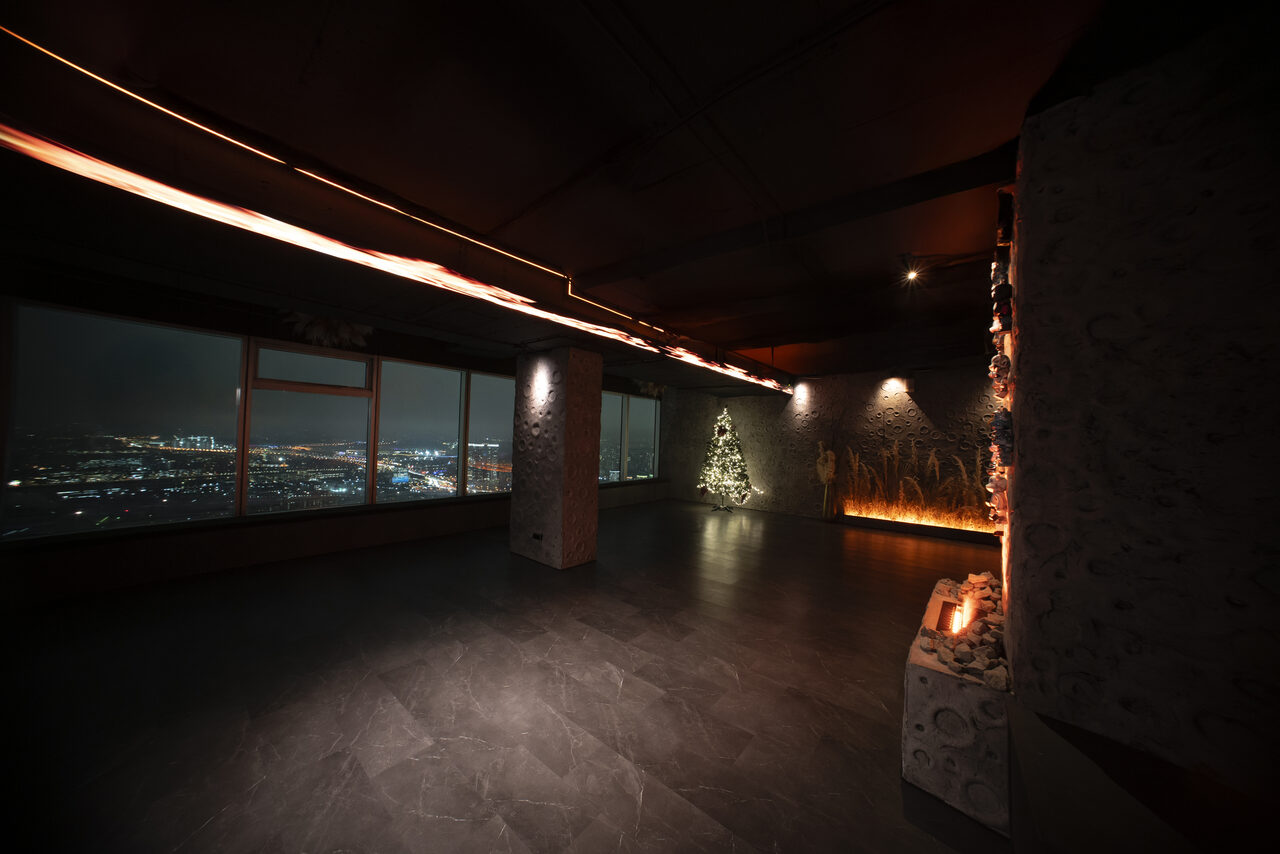 Арендовать ATMOSFERA 53(Атмосфера 53). Просторное помещение с видом на город через большие окна. Темное покрытие пола и потолка. На стене светящаяся инсталляция в виде ёлки и свечи на стойке. Освещение приглушенное, мягкое, с акцентами на элементы декора.