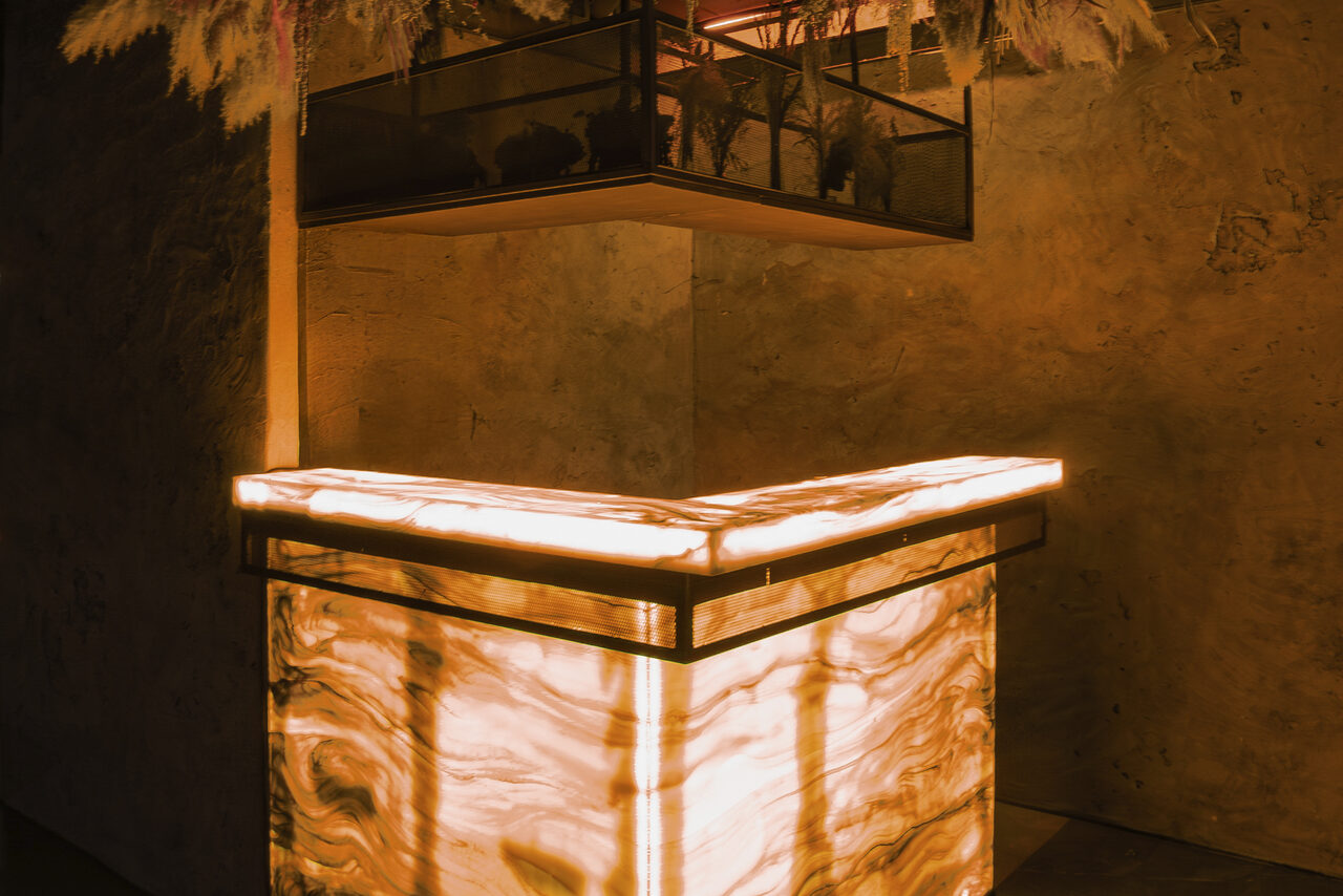 Арендовать ATMOSFERA 53(Атмосфера 53). Угловая стойка с подсветкой из полупрозрачного материала с узорами, стены с текстурой, сверху декоративные растения в подвесной конструкции, мягкое оранжевое освещение.