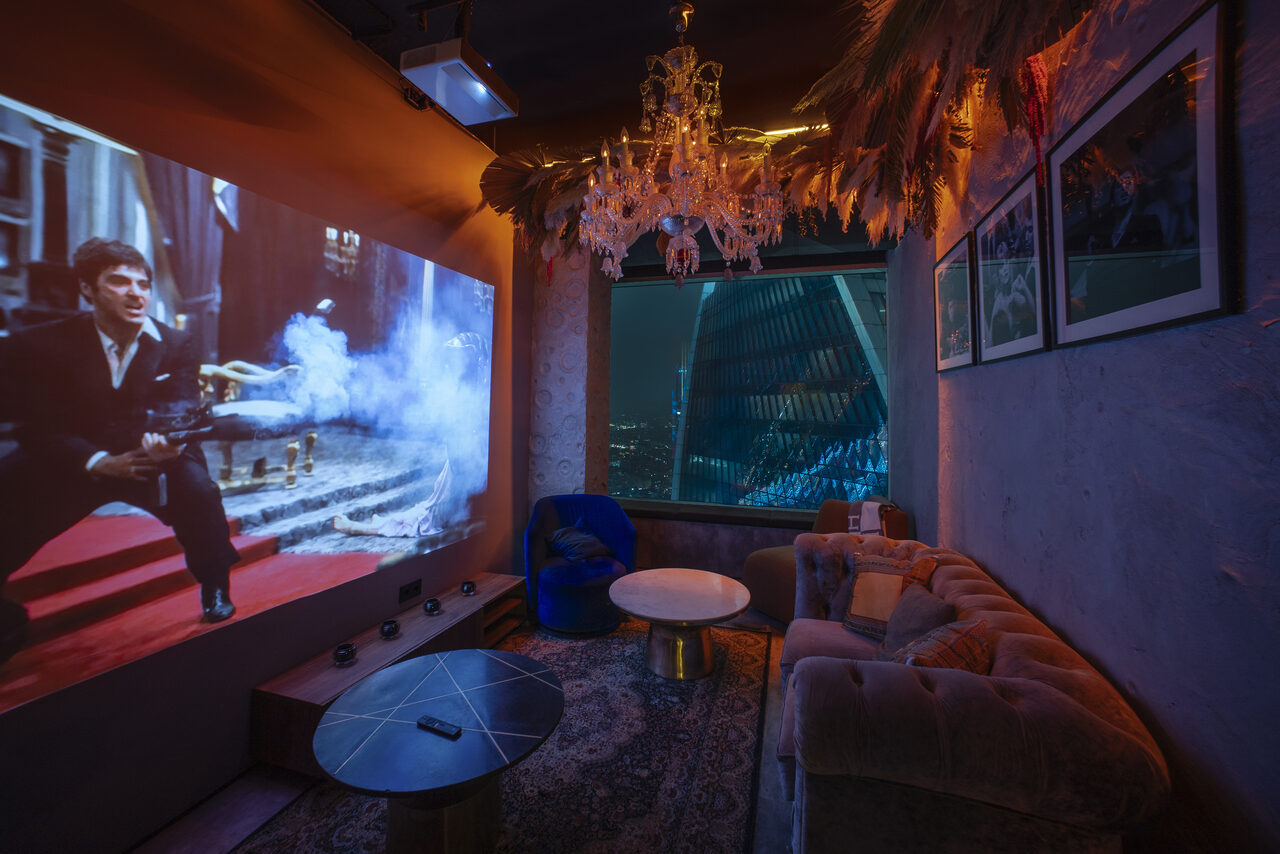 Арендовать ATMOSFERA 53(Атмосфера 53). Кинотеатр с экраном, демонстрирующим фильм. Мягкий диван, журнальные столики, ковер. Потолочная люстра, приглушенное освещение. Декоративные элементы на потолке. На стене в раме несколько картин. Вид из окна на городские здания ночью.