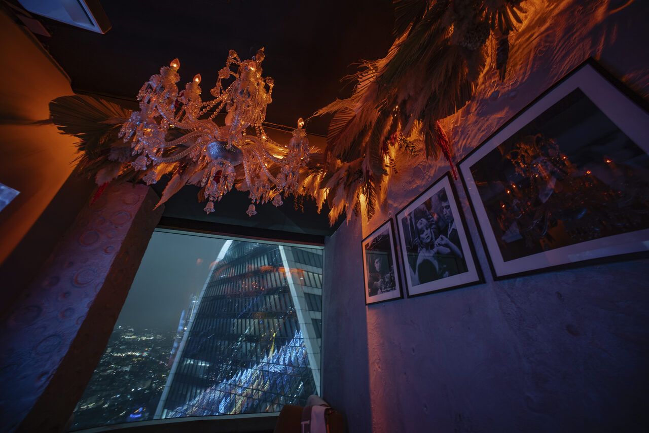 Арендовать ATMOSFERA 53(Атмосфера 53). Пространство с темными стенами и стеклянной люстрой. На стене несколько фотографий в рамках. Вид из окна на городской пейзаж с современным небоскребом. Освещение теплое, подчеркивающее декоративные элементы из растений.