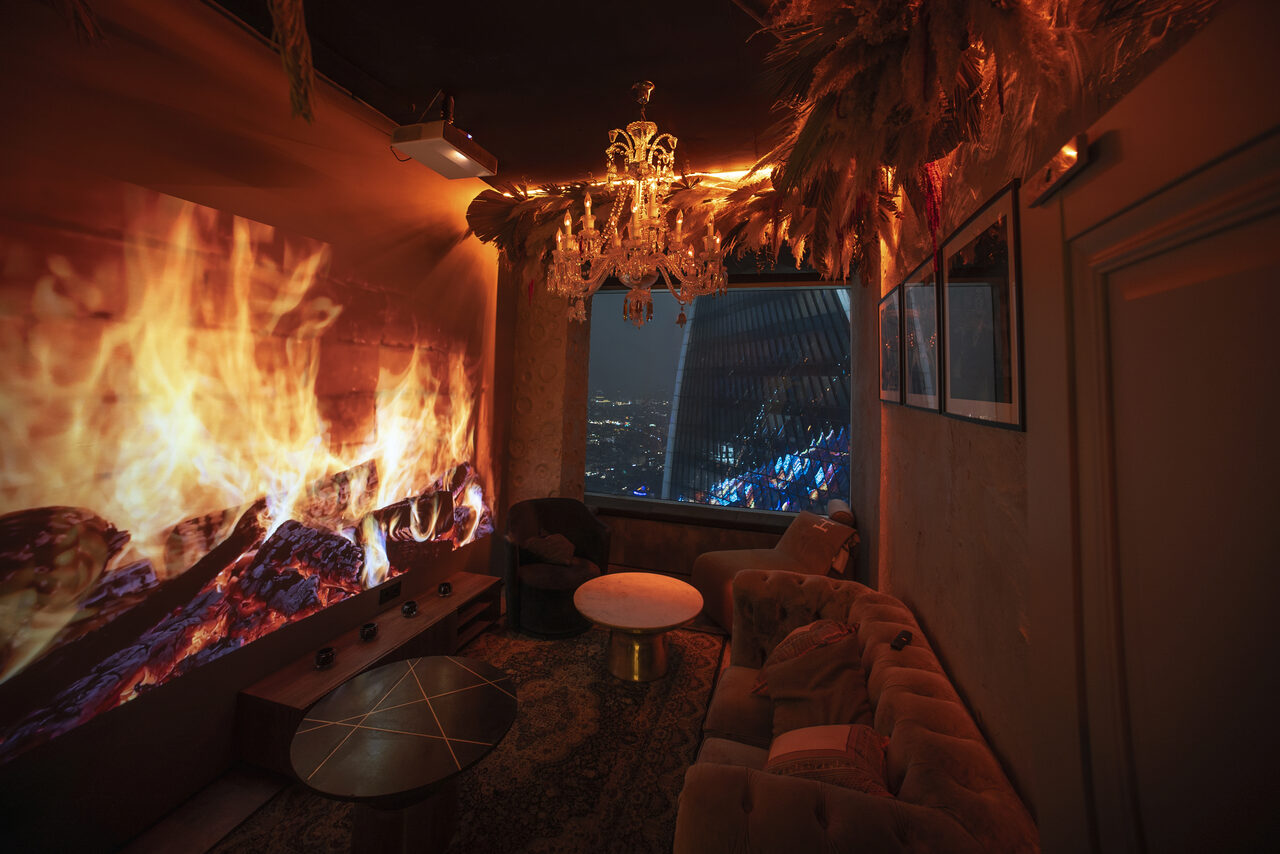 Арендовать ATMOSFERA 53(Атмосфера 53). Частное помещение с мягкой мебелью и журнальными столиками. Стена декорирована проекцией огня. Большое окно с видом на ночной город. Потолок украшен люстрой и декоративными элементами. Интерьер выполнен в тёмных теплых тонах.