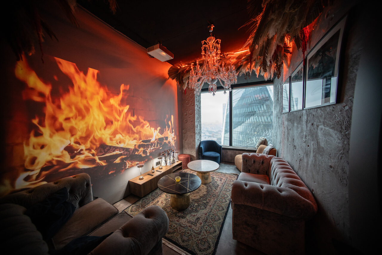 Арендовать ATMOSFERA 53(Атмосфера 53). Лофт-пространство с диванами и столами, украшенное панно с изображением огня. На потолке подвесная люстра, стены декорированы растениями. В комнате окно с видом на город, паркет и ковёр, мягкие стулья и подушки, тёплое освещение.