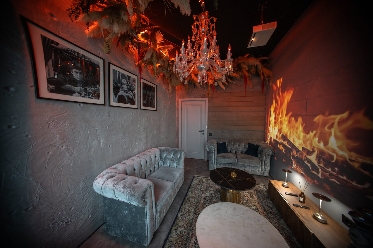 Арендовать ATMOSFERA 53(Атмосфера 53). Гостиная с серыми велюровыми диванами, на стенах фотографии в рамах и декоративные элементы. Потолочный хрустальный светильник, на стене проекция огня. На полу ковер, журнальный столик. Стены темные, полки с лампами на боку.