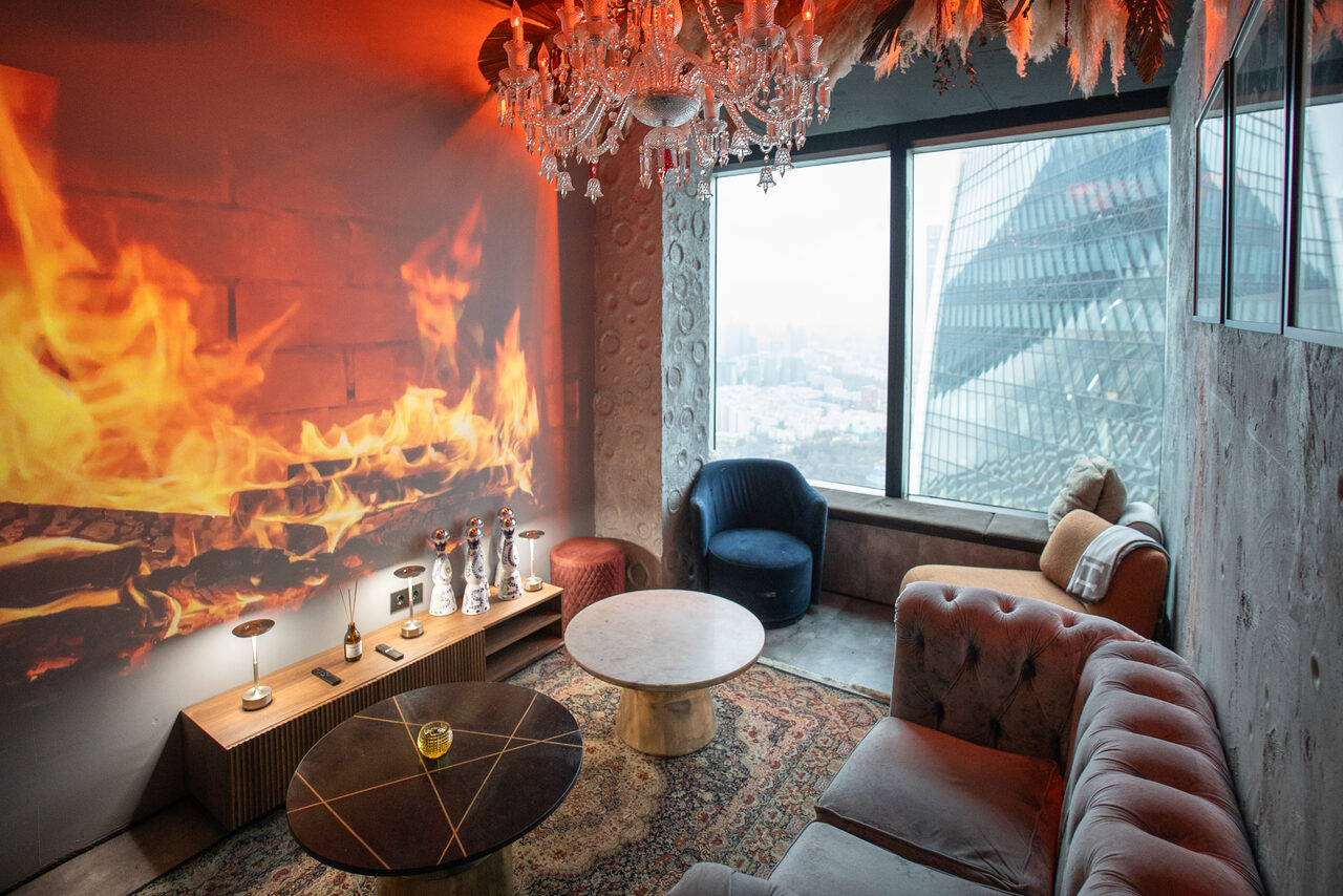 Арендовать ATMOSFERA 53(Атмосфера 53). Гостиная с панорамным окном и видом на город. Мягкие кресла и диван, два журнальных стола, декоративный камин с проекцией огня. Хрустальная люстра, настенные декорации. Стены с бежевой текстурой, ковёр с узором. Декор из статуэток и ламп на тумбе.