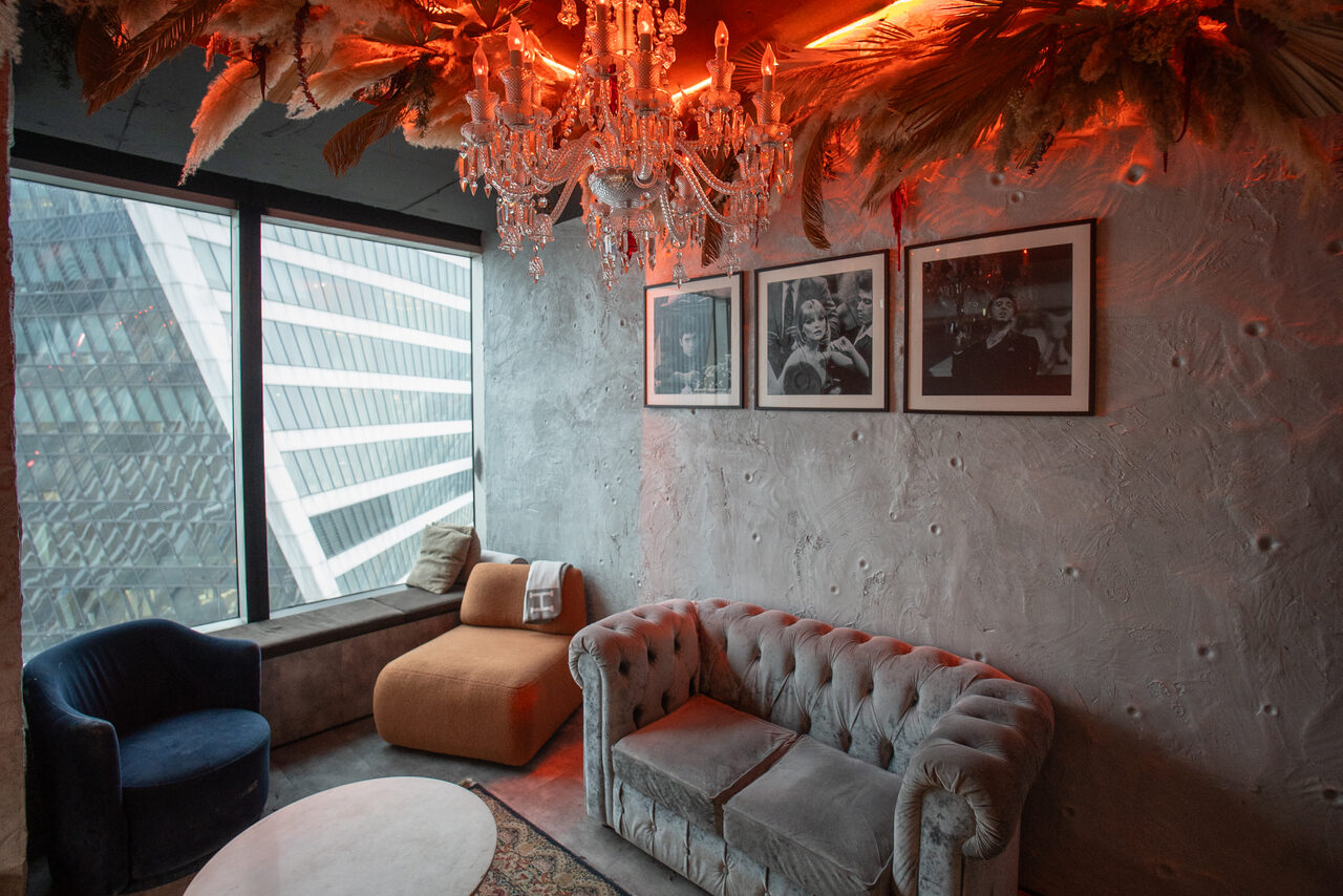 Арендовать ATMOSFERA 53(Атмосфера 53). Лофт-пространство с серыми стенами и стеклянными окнами. Мебель: серая и оранжевая мягкая мебель, круглый стол. Освещение: подвесная люстра с красной подсветкой. На стене висят три черно-белые фотографии.