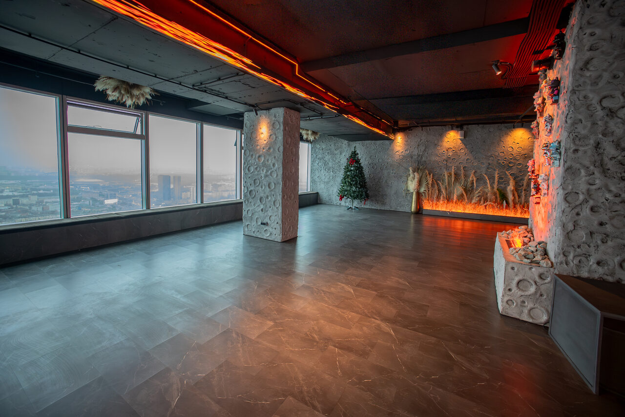Арендовать ATMOSFERA 53(Атмосфера 53). Просторное помещение с панорамными окнами и темным полом. На стенах декоративные элементы с растениями, светодиодная иллюминация. Потолок с акцентным освещением. Угловая стена оформлена камнем, присутствует елка и вазы с высокими травами.