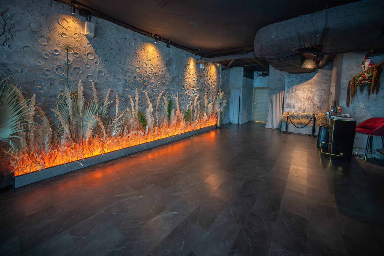 Арендовать ATMOSFERA 53(Атмосфера 53). Просторная комната с серыми стенами и полом, декоративная стена с растениями и подсветкой, потолок с вентиляцией. В правом углу стол, красный стул и цветочная композиция. Освещение точечное.