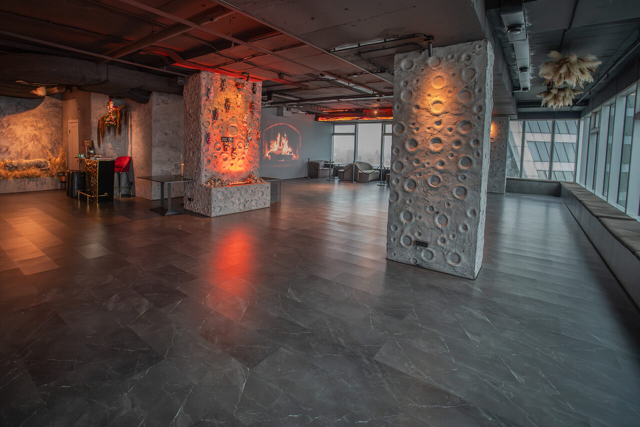 Арендовать ATMOSFERA 53(Атмосфера 53). Просторное помещение с колоннами, облицованными узорчатым материалом. Пол темный, глянцевый. На стенах проекция с изображением. Большие окна, скамьи вдоль них. Несколько зон с диванами и столами. Потолок открытый, индустриального стиля.