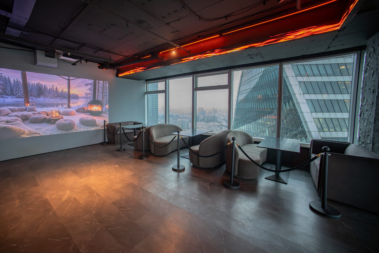 Арендовать ATMOSFERA 53(Атмосфера 53). Просторное помещение с окнами на всю высоту стены, с видом на город. Справа стоят круглые кресла и прямоугольные столики, разделенные барьерами. На стене слева проектируется изображение зимнего пейзажа. Подвесное освещение, темный пол, бетонный потолок.