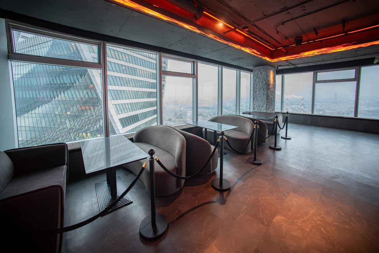 Арендовать ATMOSFERA 53(Атмосфера 53). Зал с окнами от пола до потолка, вид на городские здания. Вдоль окон — мягкие серые кресла, напротив них — столы. Потолок и стены серые, подвесное освещение с оранжевыми лампами. Пол большой и свободный, отделан под мрамор.