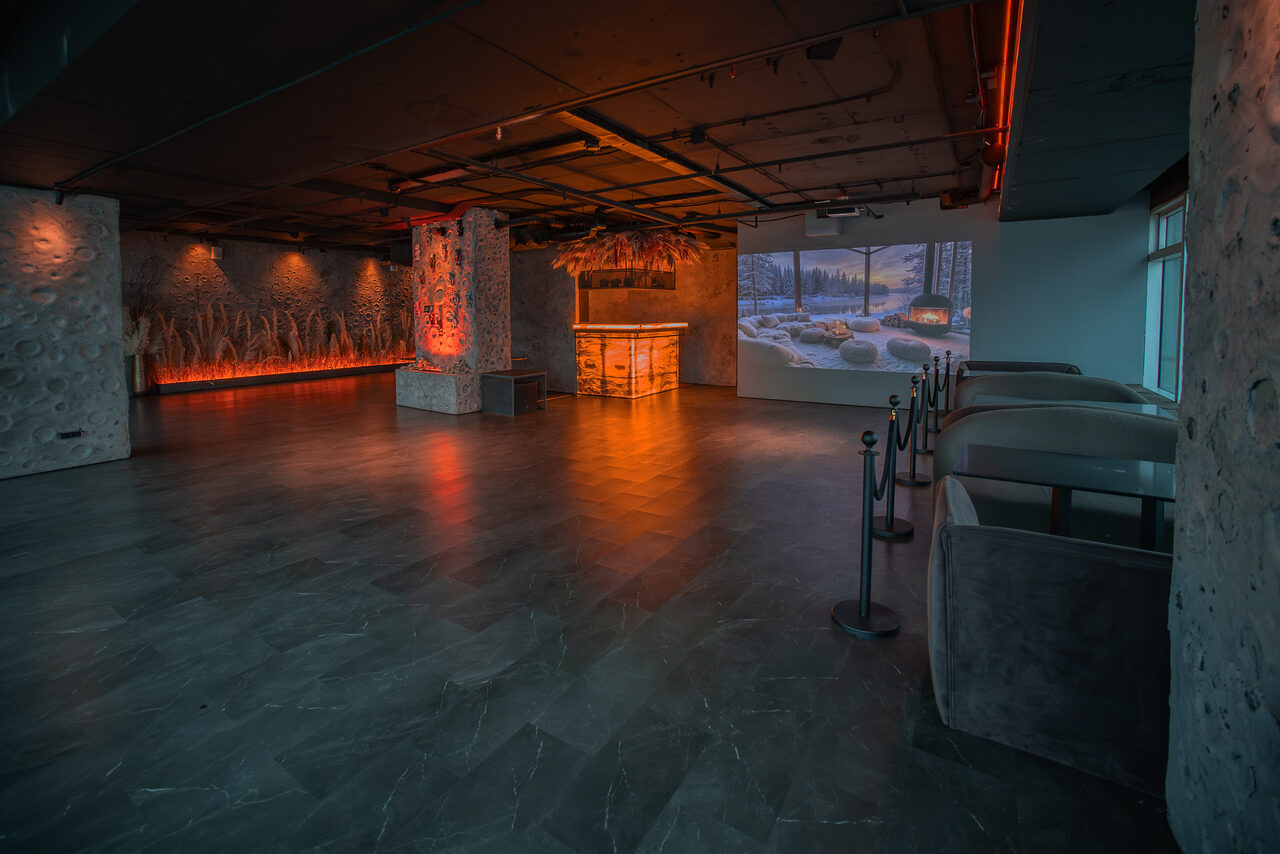 Арендовать ATMOSFERA 53(Атмосфера 53). Пространство открытой планировки с бетонным потолком, каменными и текстурированными стенами. Пол из серого камня. Мягкая зона справа с диванами и ограждением. В центре — барная стойка с подсветкой, проекция на стене, оранжевое освещение.