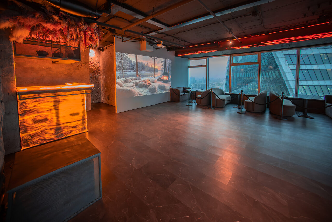 Арендовать ATMOSFERA 53(Атмосфера 53). Просторное помещение с большими окнами и видом на город. Слева – барная стойка с подсветкой. У стены – проектор с изображением зимнего пейзажа. Справа несколько кресел и столиков. Потолок с открытыми коммуникациями. Освещение тёплое, рассеянное.