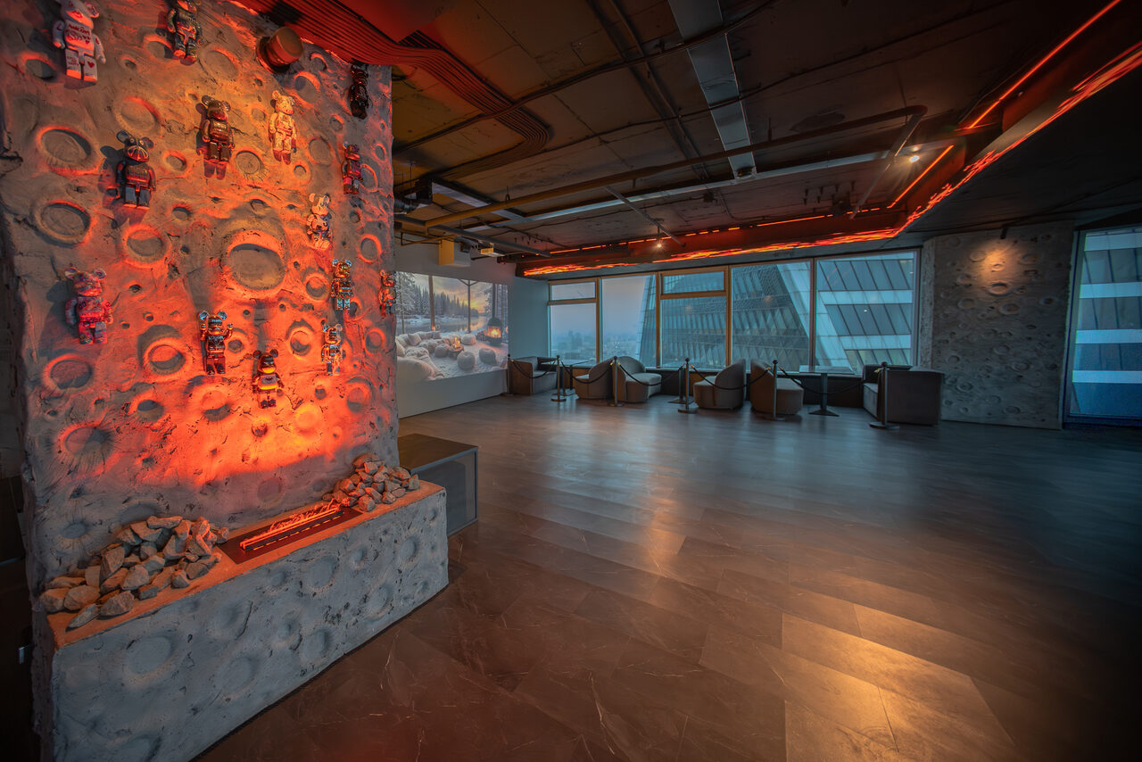 Арендовать ATMOSFERA 53(Атмосфера 53). Просторное помещение с зоной отдыха; стены и потолок индустриального стиля с коммуникациями; декоративная стена с подсветкой и фигурками; панорамные окна с видом на здания; зона отдыха с креслами; пол из темного дерева; теплое освещение.