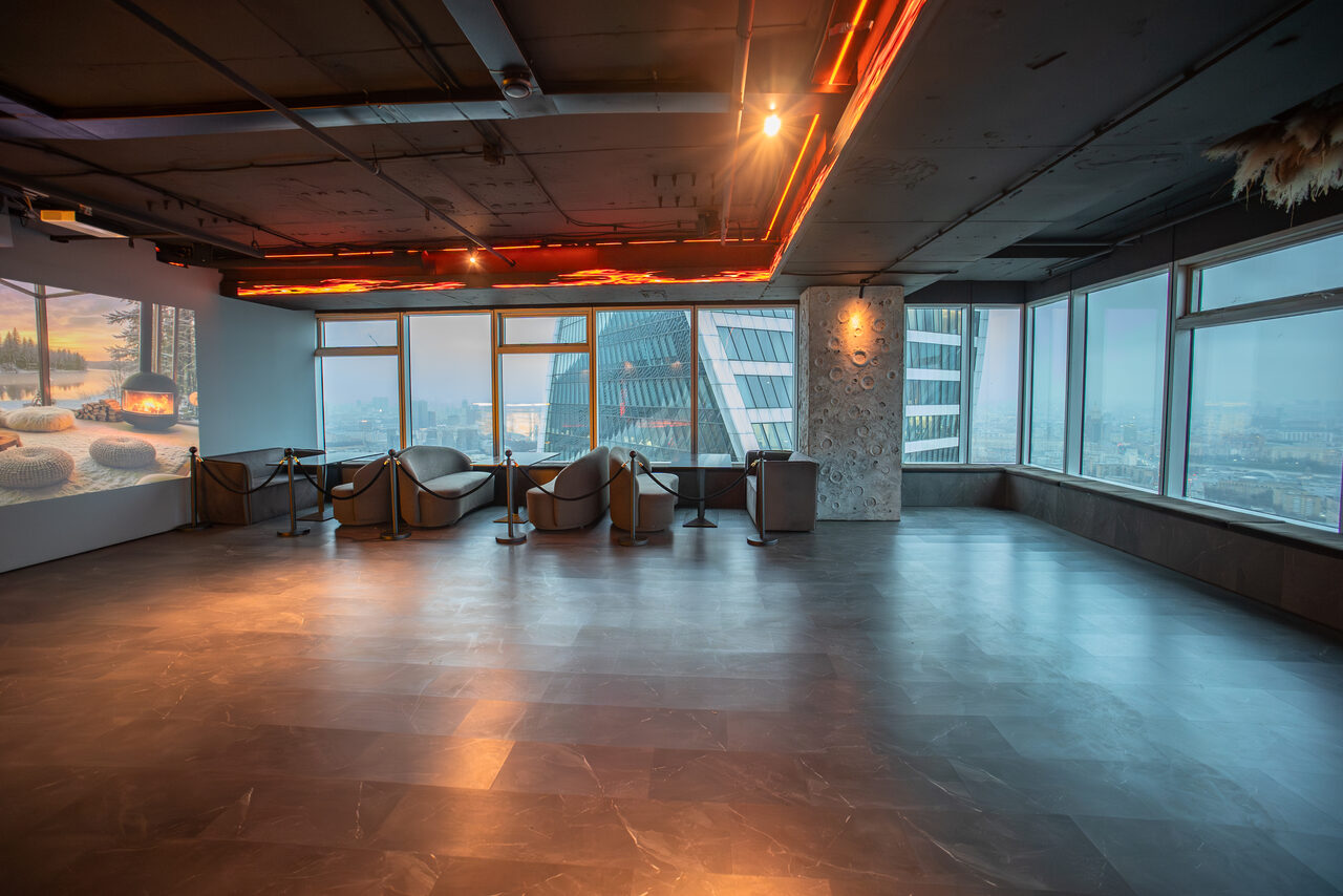 Арендовать ATMOSFERA 53(Атмосфера 53). Просторное помещение с панорамными окнами и видом на город. Темный пол, серые стены, потолок с декоративной подсветкой. Несколько мягких кресел и столов у окна. На стене проекция изображения горящего камина.