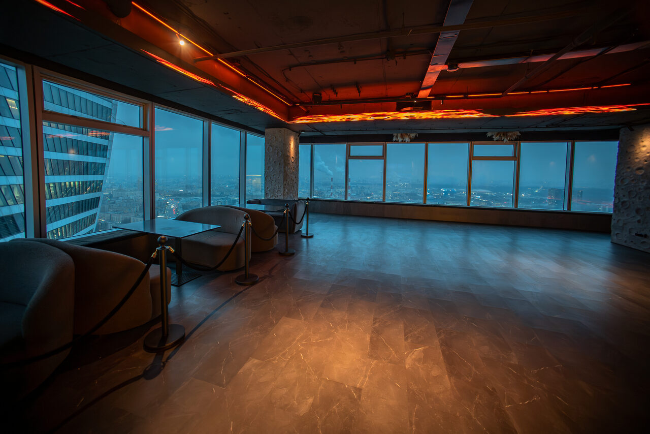 Арендовать ATMOSFERA 53(Атмосфера 53). Просторное помещение с панорамными окнами, темный пол, бетонный потолок с ярким оранжевым освещением по периметру. Небольшие столы и кресла, разделенные канатами. Вид на город. Современный стиль, открытая планировка.