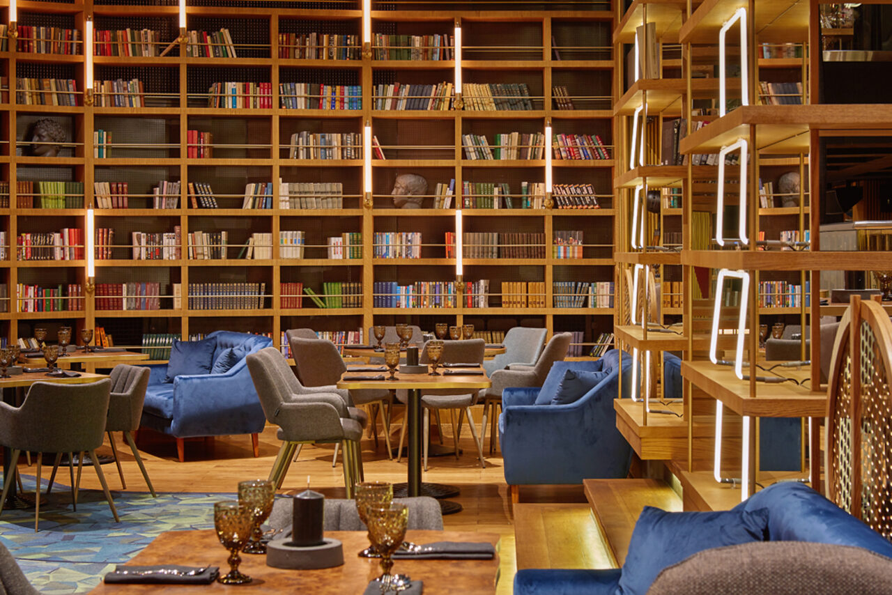 Арендовать Communitymoscow (Коммьюнити Москоу).Зал Библиотека. Многофкнкциональное пространство. Библиотечное пространство с высокой деревянной полкой, заполненной книгами. В центре расставлены круглые столы и серо-синие кресла. Потолок и пол оформлены деревянными элементами. Освещение мягкое, с акцентными светильниками.