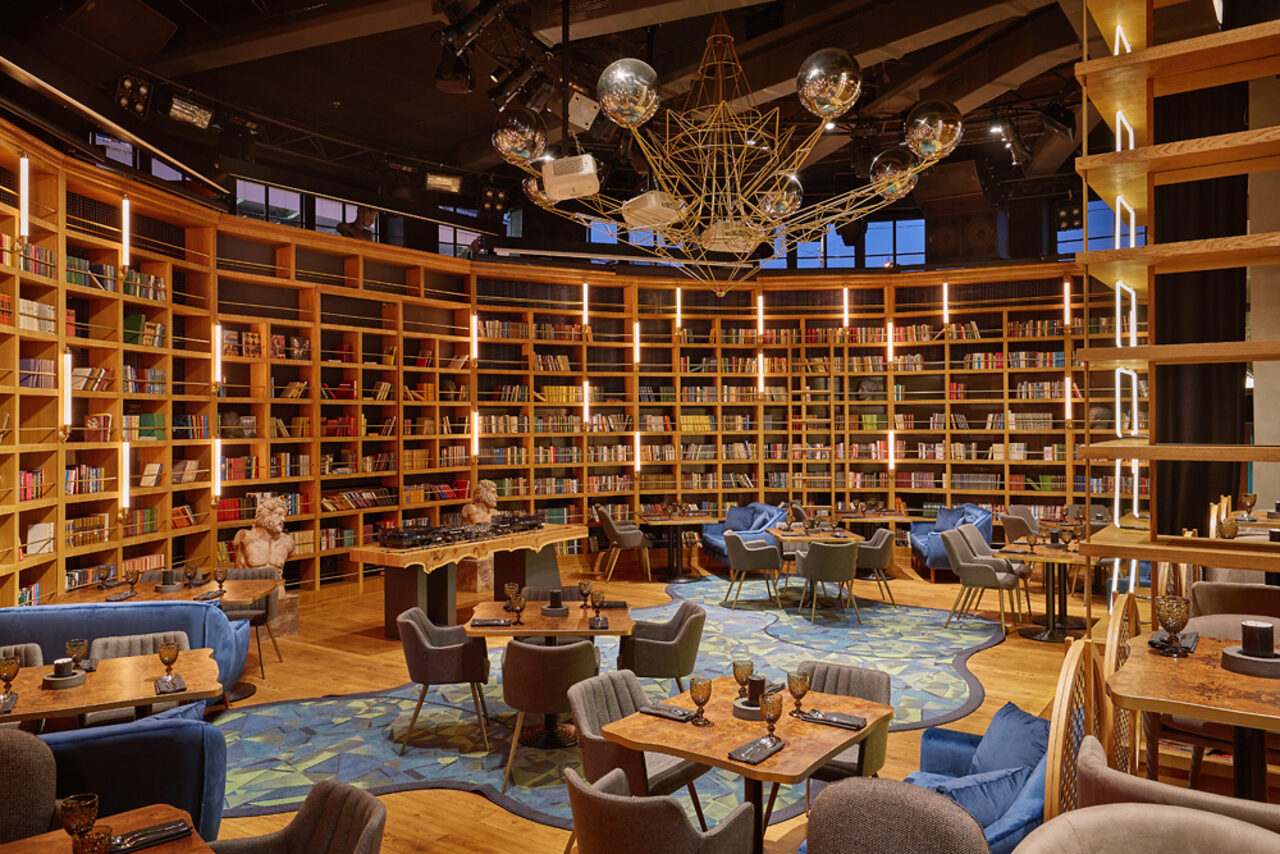 Арендовать Communitymoscow (Коммьюнити Москоу).Зал Библиотека. Многофкнкциональное пространство. Библиотечное пространство с деревянными стеллажами, заполненными книгами. Планировка с креслами и столами для отдыха и работы. Темный потолок с современными светильниками. Пол с узорчатым ковром. Приятное теплое освещение, на стенах лампы.