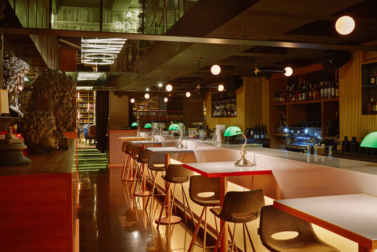 Арендовать Communitymoscow (Коммьюнити Москоу).Зал Библиотека. Многофкнкциональное пространство. Барное пространство с длинной стойкой и высокими стульями, полки с напитками на стене. Подвесные круглые светильники, настольные лампы с зелеными абажурами. Темное деревянное и металлическое оформление, зеркальные потолки и светящаяся надпись.