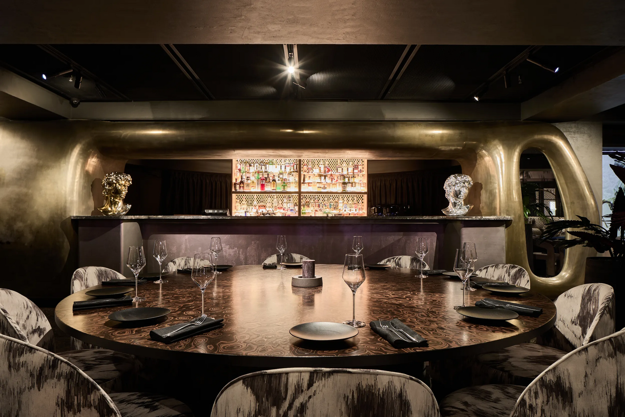 Арендовать Communitymoscow (Коммьюнити Москоу).Зал Библиотека. Многофкнкциональное пространство. Круглый стол с несколькими стульями, сервированный пустыми тарелками и бокалами. На заднем плане барная стойка с полками для бутылок. Потолочное освещение, стены и бар отделаны в золотистых тонах. Темное общее освещение, современный интерьер.