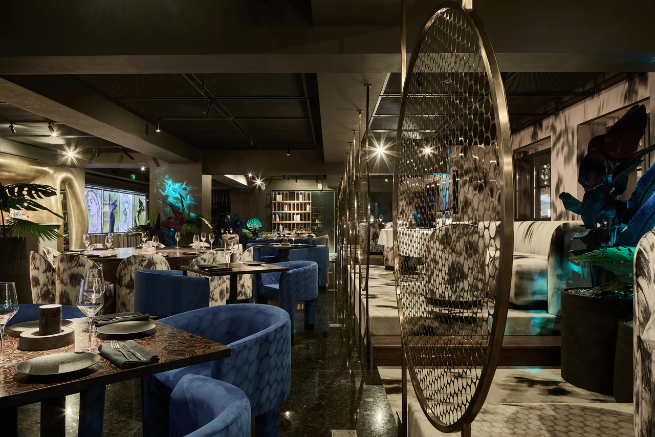 Арендовать Communitymoscow (Коммьюнити Москоу).Зал Библиотека. Многофкнкциональное пространство. Ресторанное пространство с круглым деревянным столами и синими креслами. Металлические перегородки, полированное темное напольное покрытие. Настенные декоративные элементы, направленное освещение на потолке, зеленые растения, мягкие диваны в интерьере.