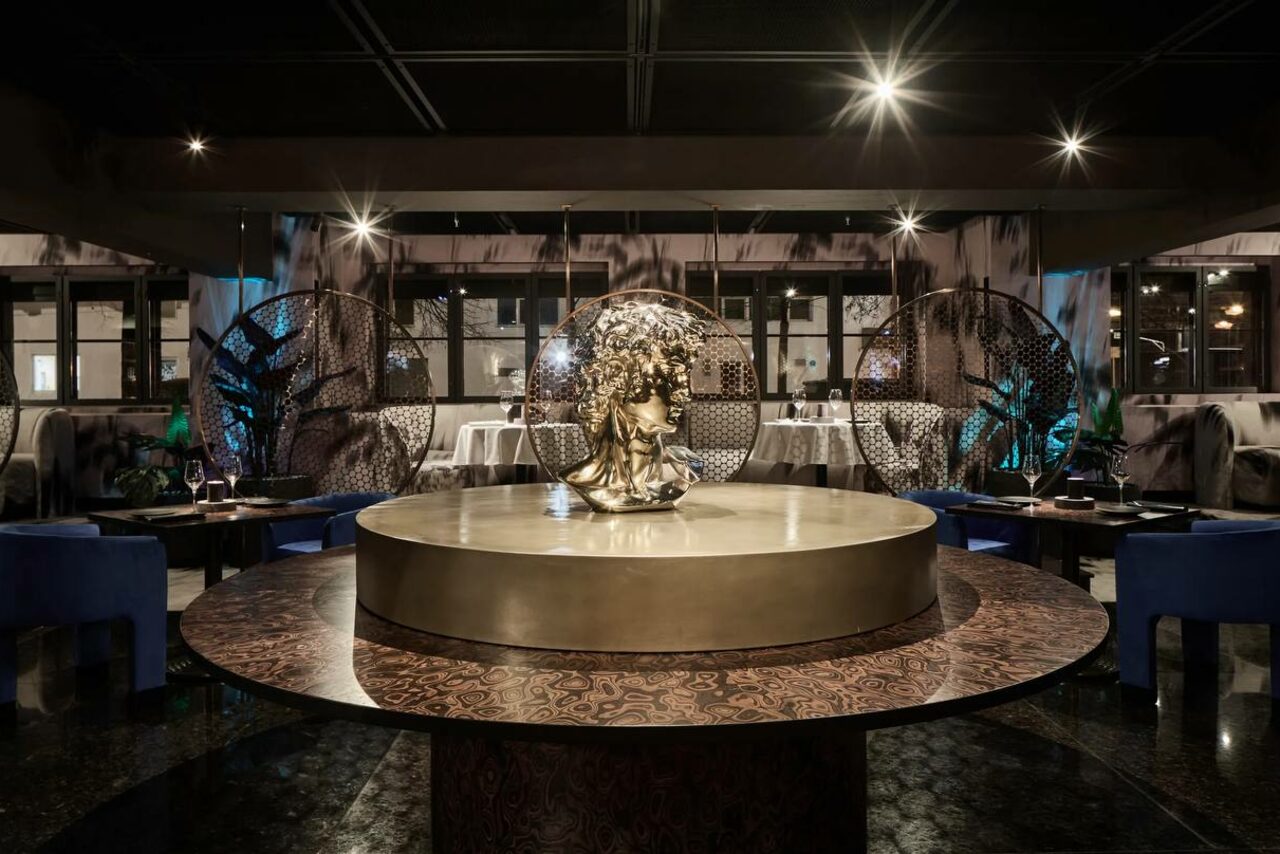 Арендовать Communitymoscow (Коммьюнити Москоу).Зал Библиотека. Многофкнкциональное пространство. Ресторанное пространство с круглым столом в центре, украшенным скульптурой головы. Мягкие кресла темно-синего цвета, круглые декоративные перегородки, черный пол, приглушенное освещение, темные стены с рисунками, окна на заднем плане.