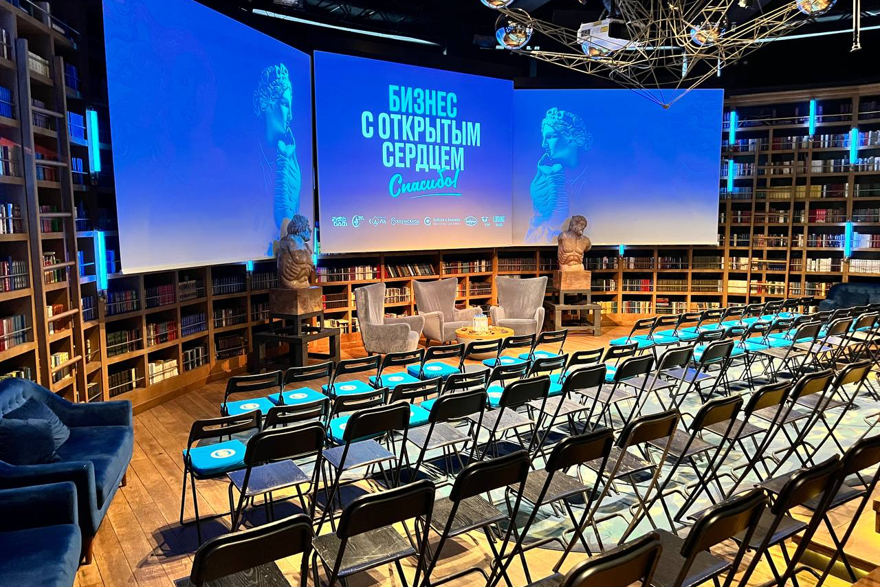 Арендовать Communitymoscow (Коммьюнити Москоу).Зал Библиотека. Многофкнкциональное пространство. Пространство для мероприятий с амфитеатром, стульями в ряд. Деревянный пол, полки с книгами вдоль стен. Три экрана с изображением, надписи на русском языке: «Бизнес с открытым сердцем. Спасибо!». Скульптуры по бокам сцены, два кресла и столик.