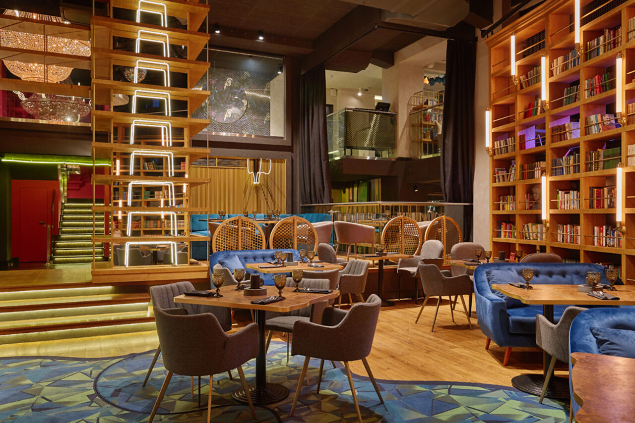 Арендовать Communitymoscow (Коммьюнити Москоу).Зал Библиотека. Многофкнкциональное пространство. Современное пространство с деревянными полами, полками с книгами, мягкими креслами и диванами. На полу узорчатый ковер, есть круглые обеденные столы. Освещение из встроенных светильников. Декор в теплых тонах.