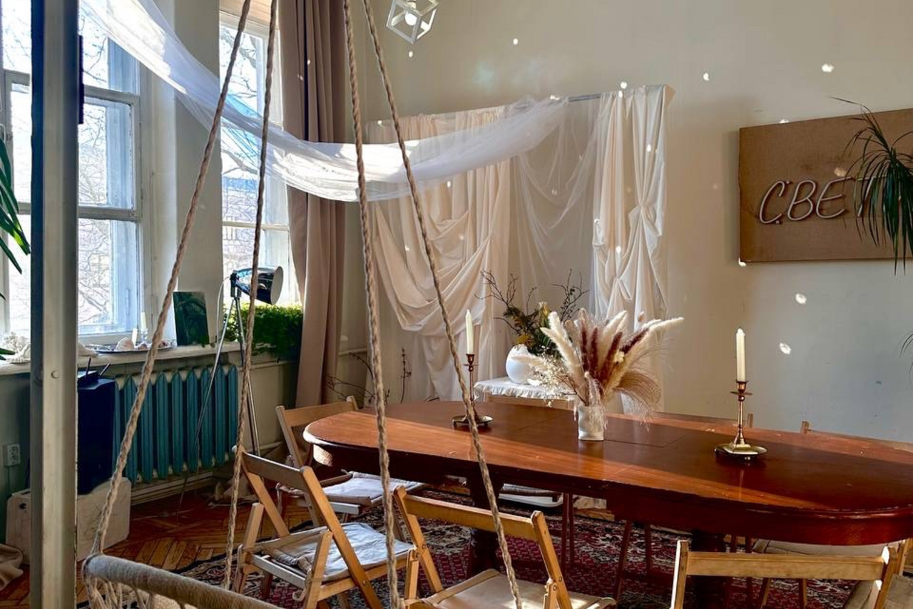 Арендовать Студия Свети. Пространство с деревянным столом, окружённым складными стульями. На столе свечи и ваза с сухоцветами. Светлая ткань подвешена у окна и на стене. Окно с занавесками, синий радиатор. На стене знак "СВЕТ". Паркетный пол, ковёр.