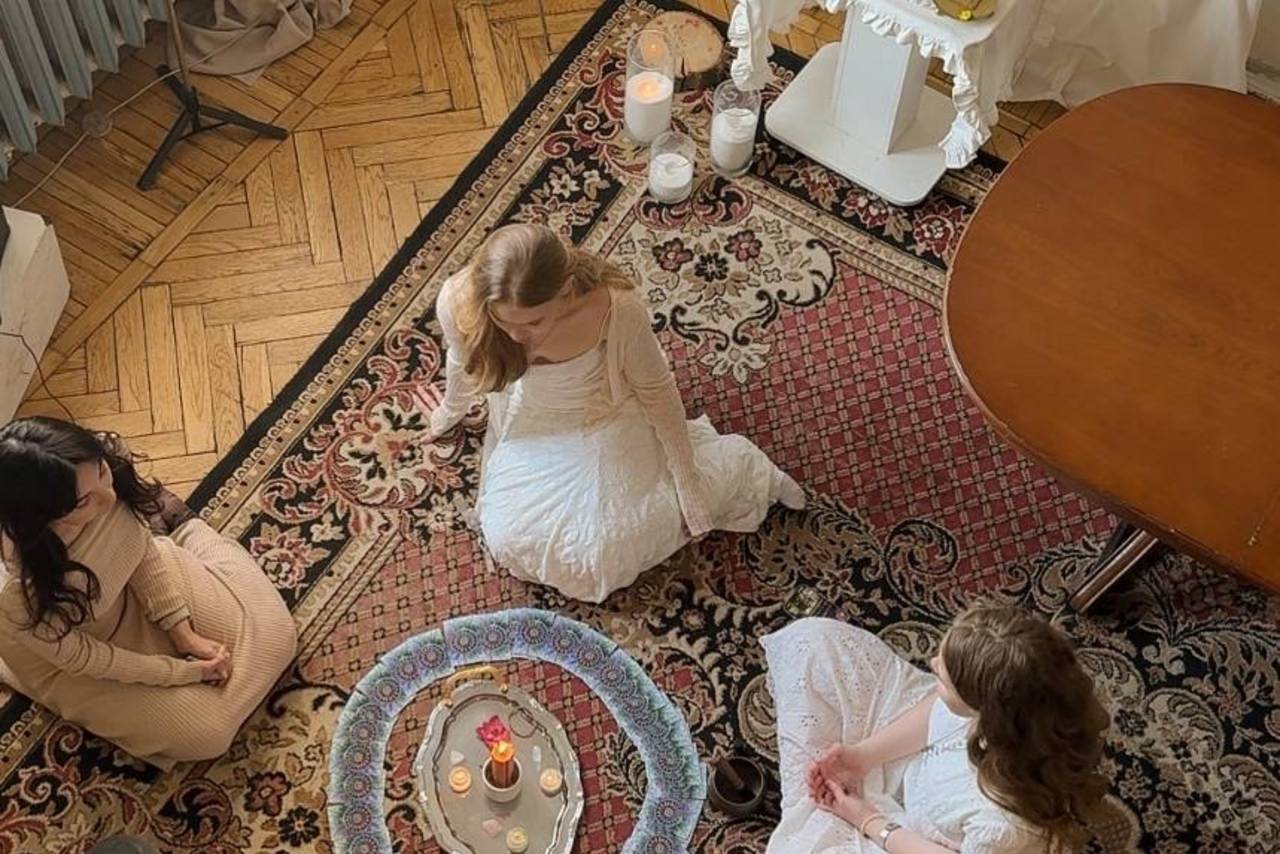 Арендовать Студия Свети. Пространство с паркетным полом и ковром с узором, круглый столик с декоративными свечами. Три человека сидят на ковре. Светлые и темные тона в интерьере, свечи добавляют мягкое освещение.