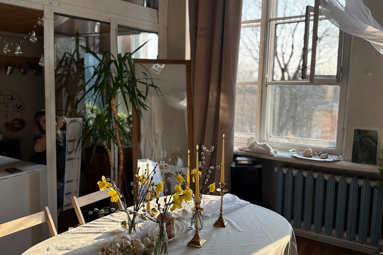 Арендовать Студия Свети. Просторная комната с круглым столом, покрытым белой скатертью, в центре цветочные декорации и золотые подсвечники. Возле окна с бежевыми шторами радиатор отопления. Зеркало и крупное растение в углу. Светлый и уютный интерьер.