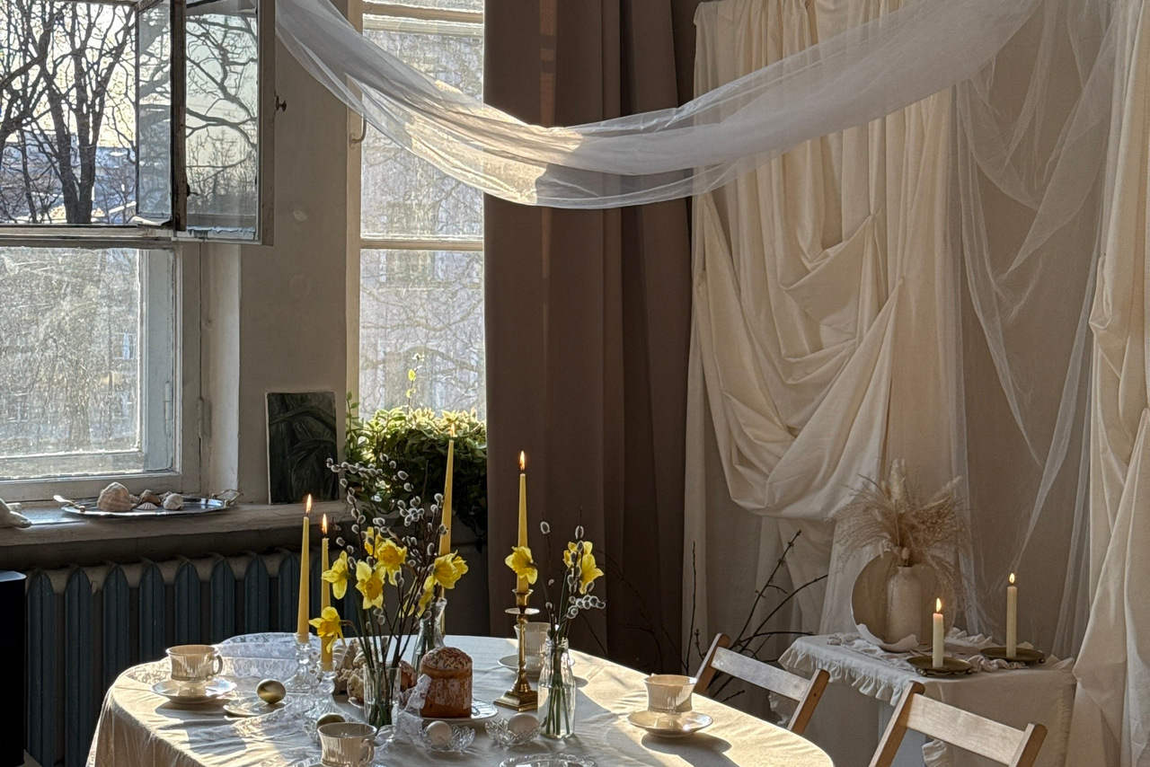 Арендовать Студия Свети. Просторная комната с большими окнами и открытыми занавесками. В центре стола белая скатерть, на которой расположены свечи, цветы и кофейные чашки. На фоне драпировки из белой ткани. Естественное освещение, дерево и текстиль в декоре.