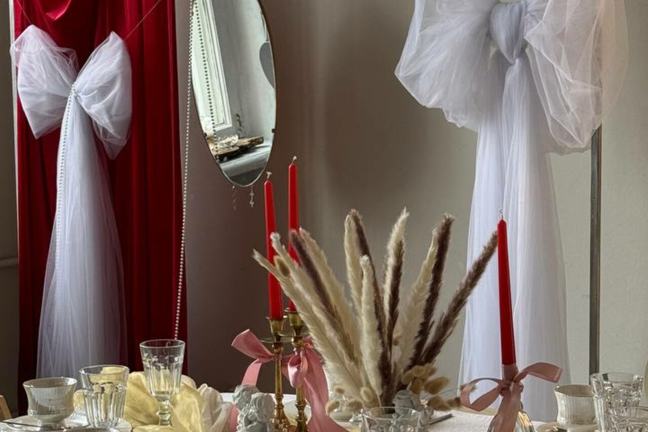 Арендовать Студия Свети. Столовая с круглым столом, на котором расположены украшения из сухоцветов, свечи в подсвечниках и стеклянная посуда. На стенах красные и белые занавеси. За столом зеркало на стене.
