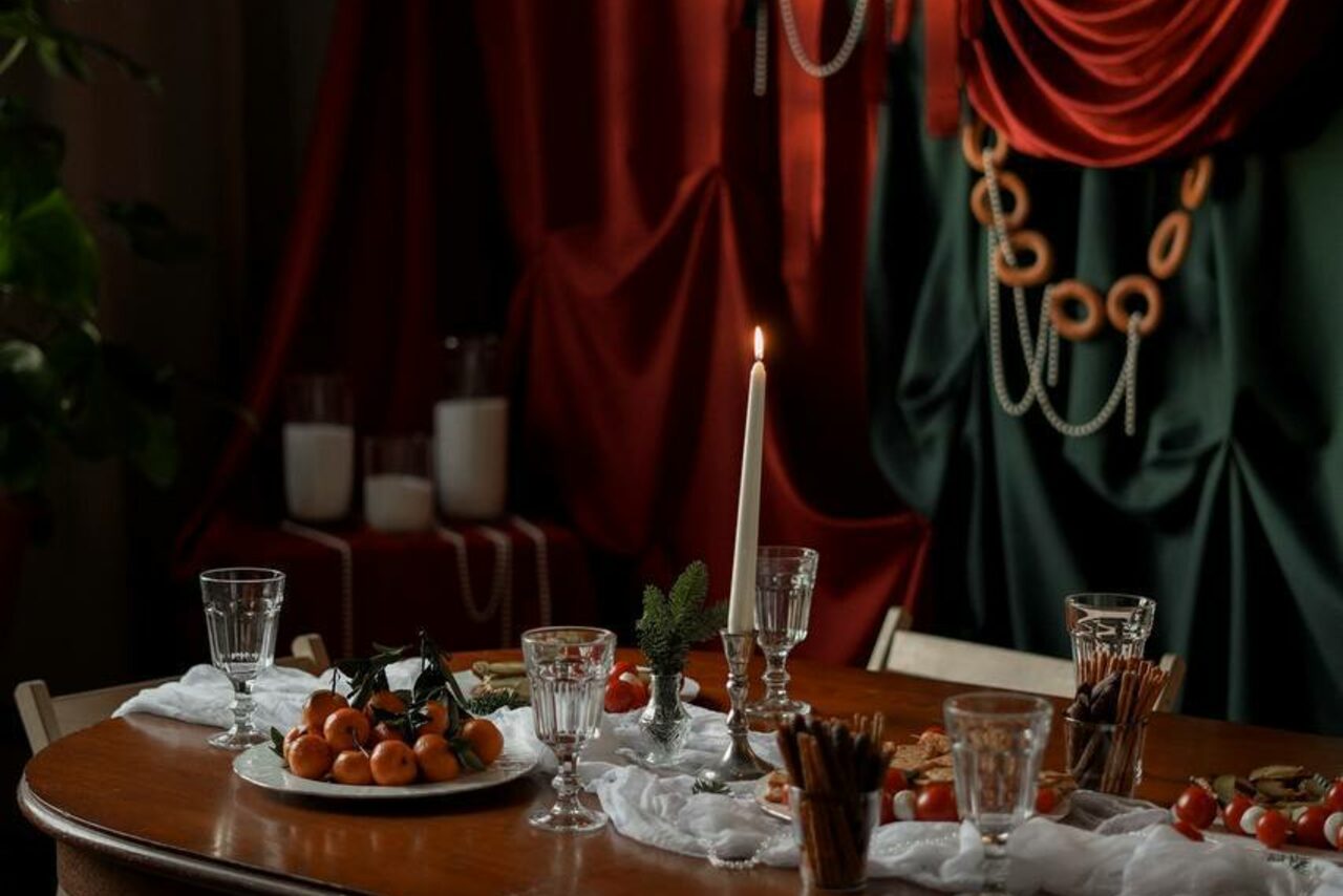 Арендовать Студия Свети. Обеденный стол с белой скатертью, украшенный фруктами и свечей. На столе расставлены бокалы, декоративные элементы из зелени. На заднем фоне темные ткани красного и зелёного цвета с гирляндами из колец и бус. Свет мягкий, атмосфера уютная.