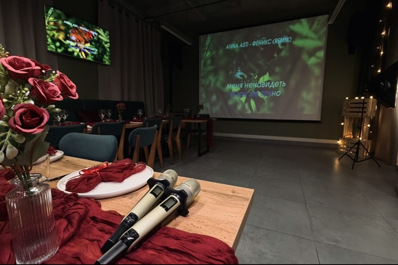Арендовать Лофт Интра. Зал Фрей. Просторное помещение с проектором, показывающим текст песни. Есть стол с красными украшениями, букетом роз и двумя микрофонами. На стене за столом висит второй экран. Стулья мягкие, темно-синие. Пол облицован плиткой. Освещение приглушенное.