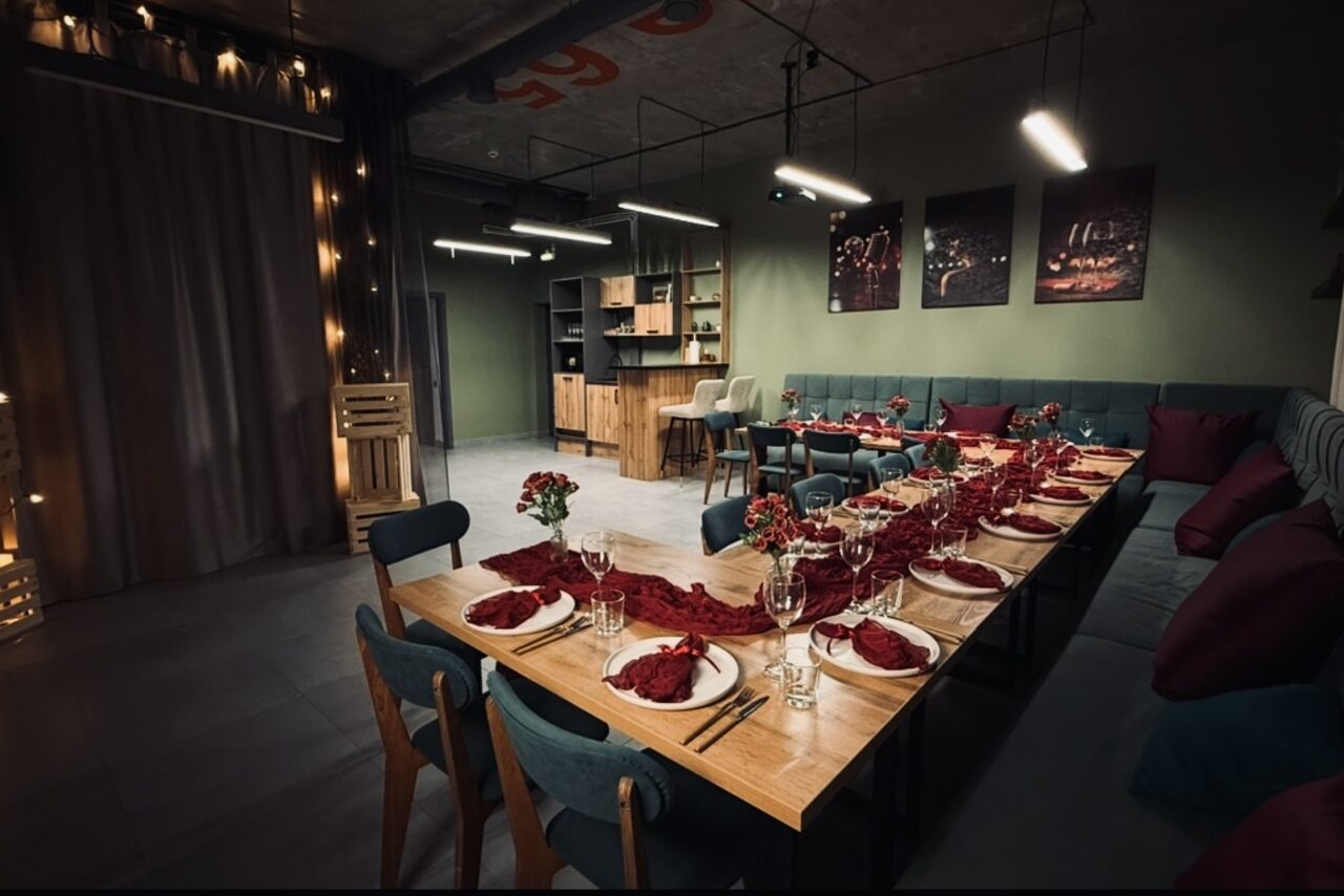 Арендовать Лофт Интра. Зал Фрей. Современное пространство с длинным деревянным столом, сервированным на десять персон, стол украшен красными салфетками и цветами. Слева занавеска, мягкий угловой диван вдоль стен, столы и деревянные стулья, открытые стеллажи. Освещение центральное и подвесное.