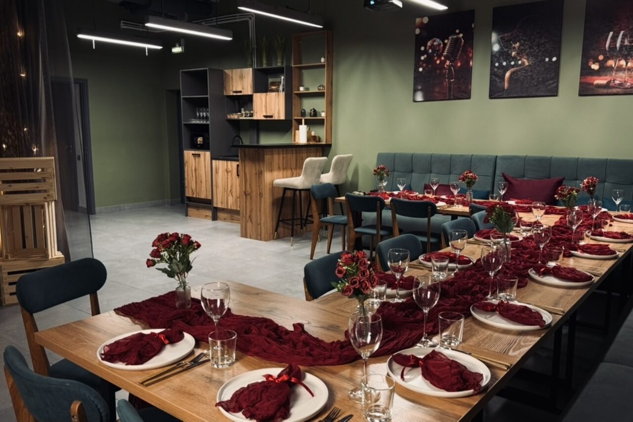 Арендовать Лофт Интра. Зал Фрей. Современное пространство с длинным столом, сервированным на 18 персон. Декорировано тканью и цветами бордового цвета. Барная стойка с высокими стульями, мягкие диваны зелёного цвета. На стене зелёного цвета висят картины. Потолочное освещение.