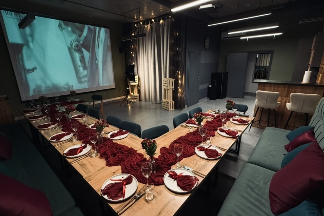 Арендовать Лофт Интра. Зал Фрей. Зал для мероприятий с экраном и проектором, U-образная расстановка столов, деревянные столы с сервировкой; красочные салфетки, бокалы и тарелки. Мягкие диваны и стулья. Подвесные светильники, нейтральные тона, темные шторы, декоративные огни.