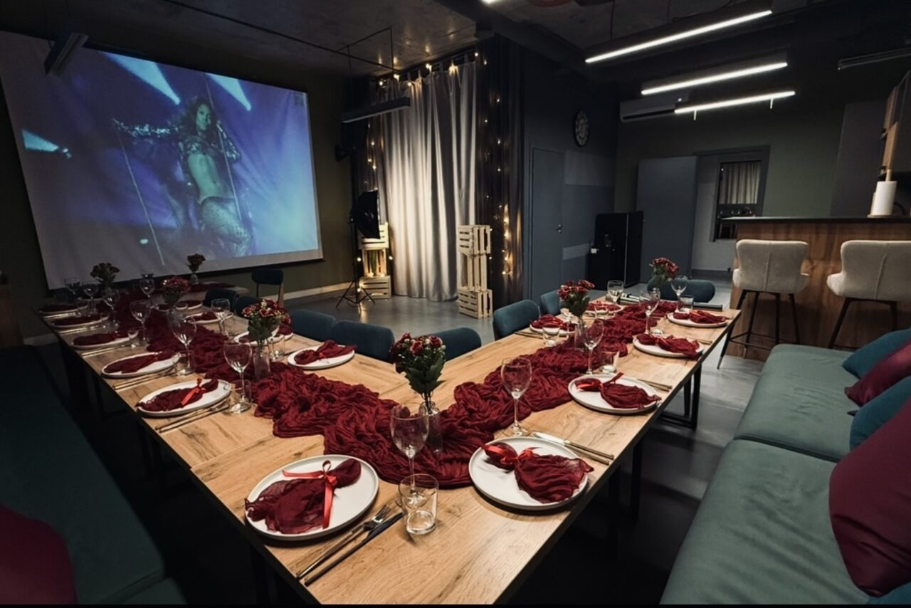 Арендовать Лофт Интра. Зал Фрей. Просторная комната с U-образным деревянным столом, сервированным красными салфетками и бокалами. На стене напротив - экран с проекцией. Серая штора, мягкие диваны, высокий барный стол, подвесные светильники. Тёмные стены и потолок.