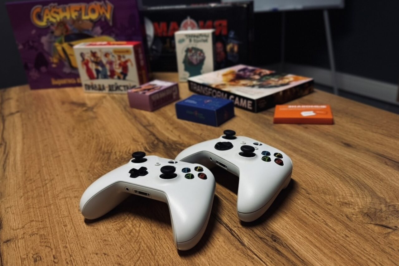 Арендовать Лофт Интра. Зал Фрей. Стол с деревянной поверхностью, на котором лежат два белых игровых контроллера. На заднем плане расположены коробки с настольными играми. Освещение приглушенное, тёмные стены на заднем фоне.