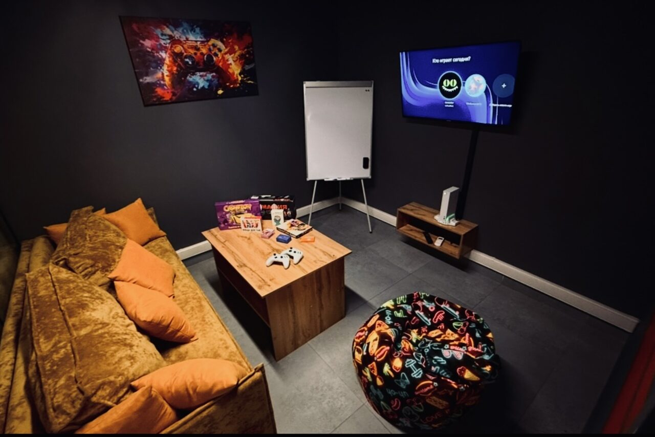 Арендовать Лофт Интра. Зал Фрей. Комната с диваном и цветными подушками, журнальный столик, пуф с ярким принтом, телевизор на стене, настольная игра, настольные принадлежности, белая доска на стенде. Темные стены, светлый пол, настенное изображение.